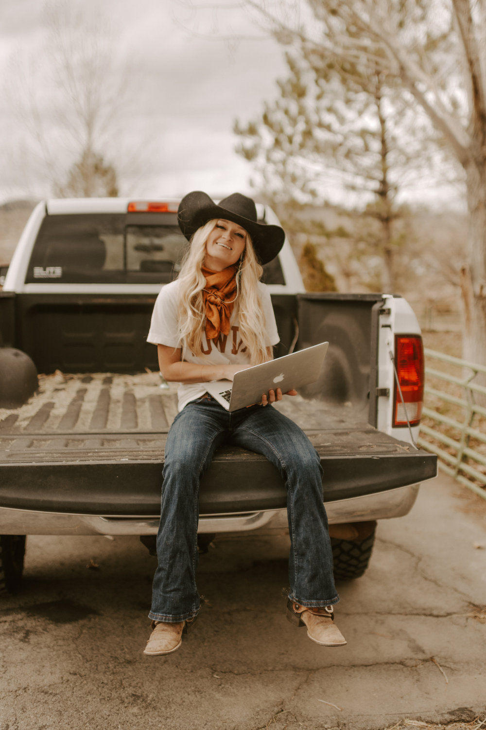 Sarah Elrod, wearing a cowboy hat, sitting on a pickup truck tailgate holding a laptop